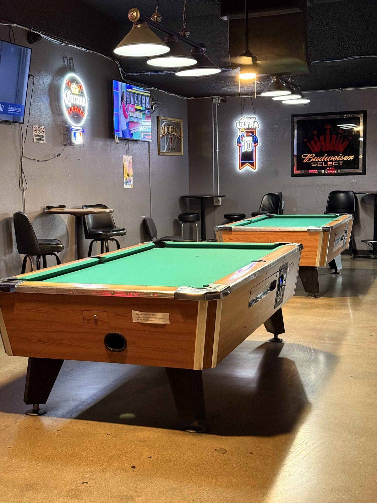 Pool room at Dingers Sports Bar. Two green-felt tables under tungsten pendant lights. Corona, IPA, and Michelob Ultra neon signs on the walls.