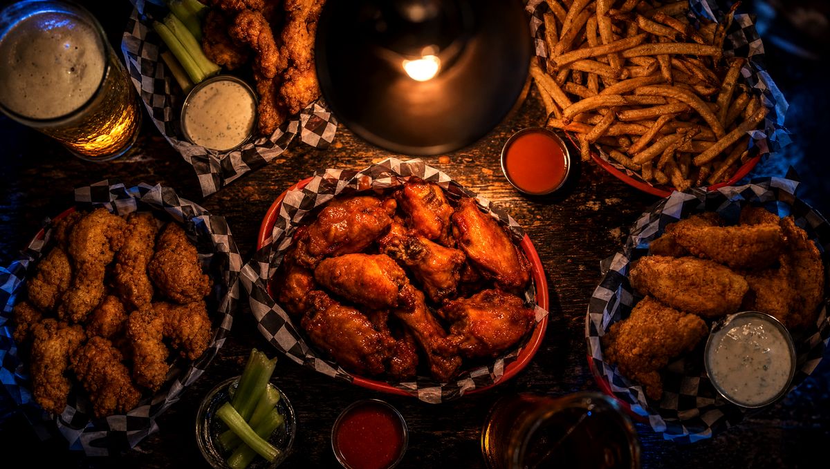 Overhead aerial shot of a sports bar food spread on dark weathered wood. Red baskets of golden chicken tenders, buffalo wings, and crispy fries. Ramekins of buffalo sauce and ranch. A pint of amber draft beer. Warm tungsten light with cool cobalt rim light.